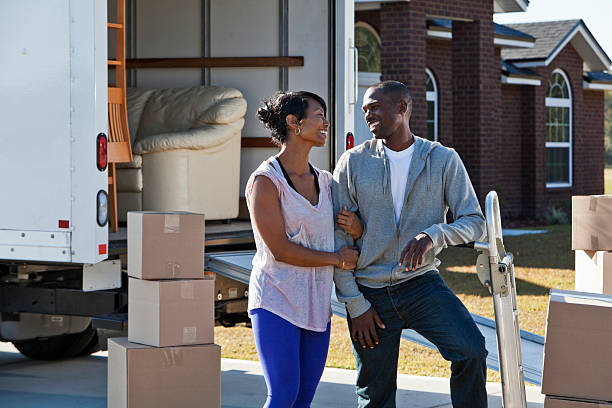 African American couple (30s) with moving truck in front of brick house, loading or unloading boxes and furniture.
